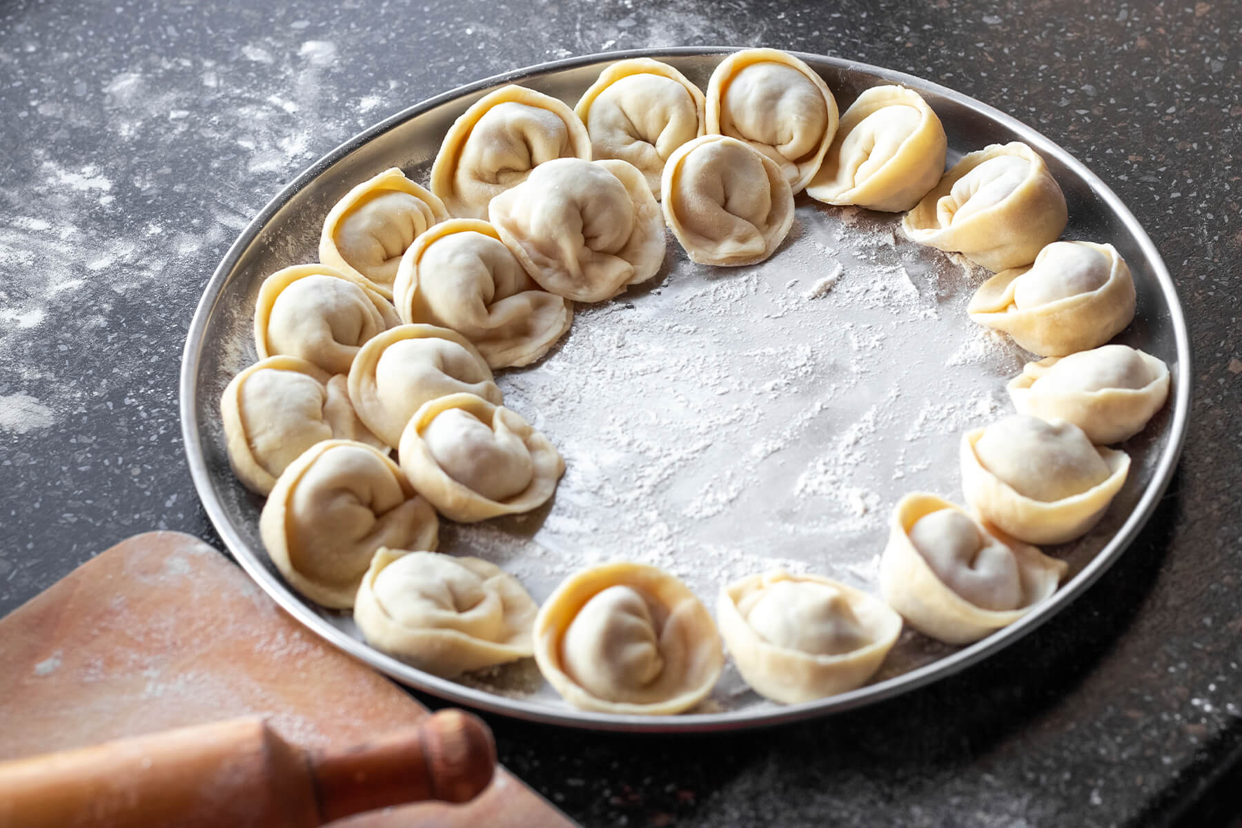 Rohe Pelmeni in einem offenen Ring mit etwas Abstand auf einem bemehlten runden Metalltablett, vorn ein Nudelholz auf einem Holzbrett