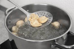 Drei gegarte Pelmeni auf einer Schaumkelle aus Edelstahl über einem Topf mit kochendem Wasser