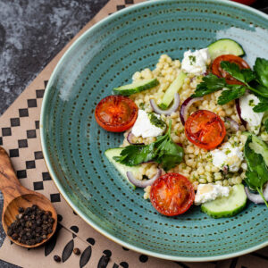 Türkisfarbener Teller mit Perlnudel-Salat, gebratenen Tomatenhälften, Gurke, roten Zwiebeln und Feta