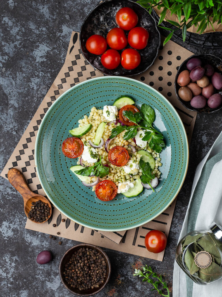 Perlnudel-Salat mit Feta, gebratenen Tomatenhälften, Gurke und Petersilie auf türkisfarbenem Teller von oben