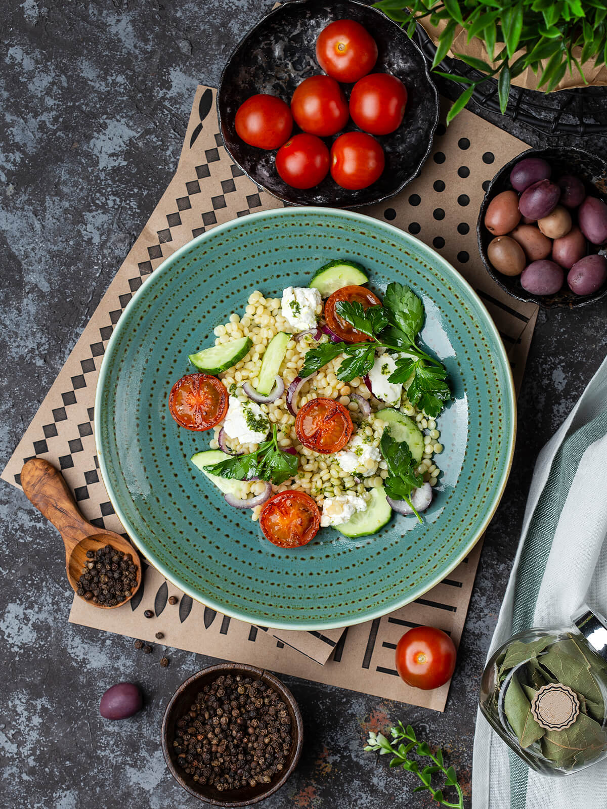 Perlnudel-Salat mit Feta, gebratenen Tomatenhälften, Gurke und Petersilie auf türkisfarbenem Teller von oben