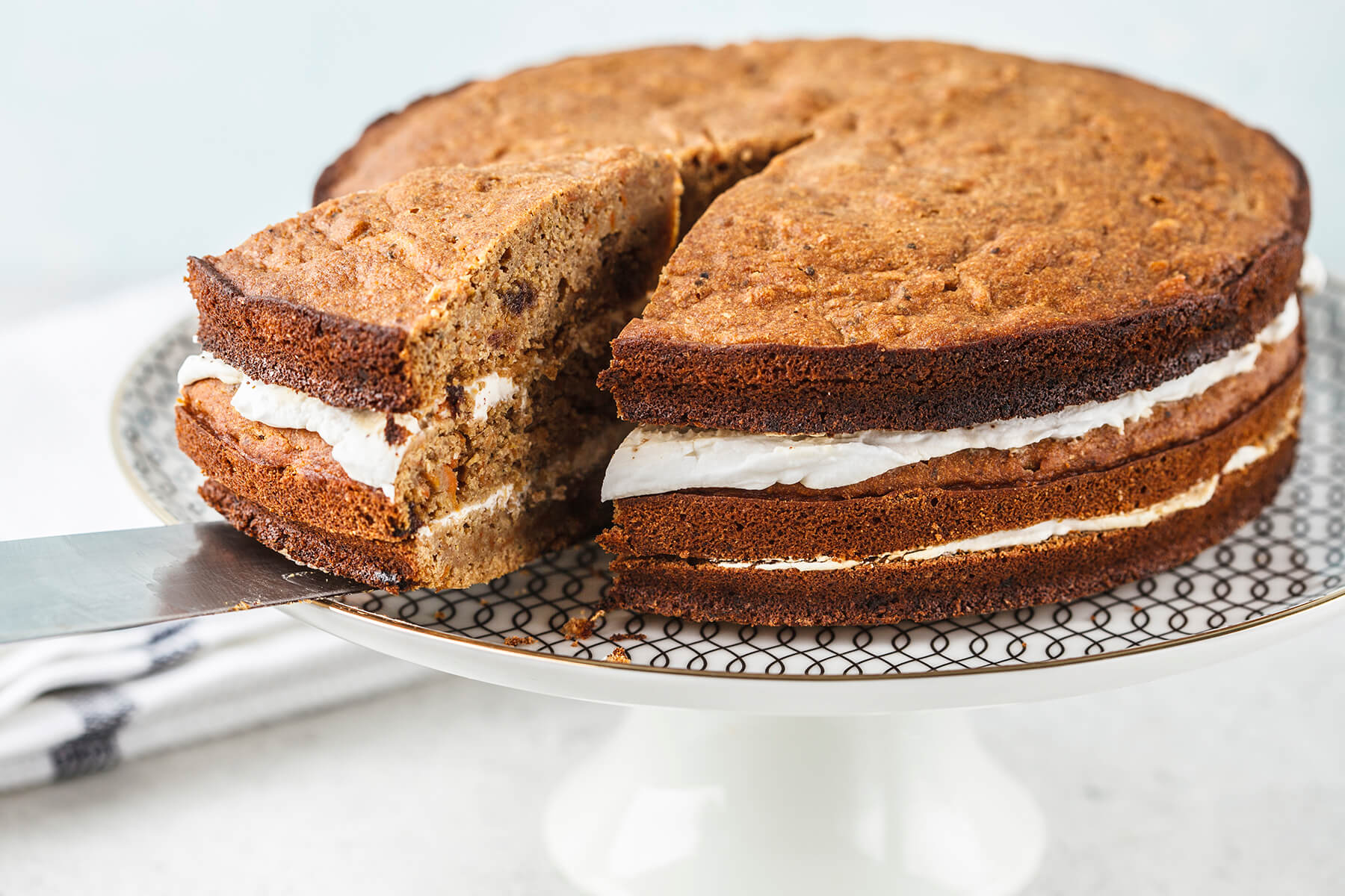 Veganer Karottenkuchen aus drei Vollkornböden mit zwei Schichten Kokoscreme, ein Stück wird mit dem Messer abgehoben