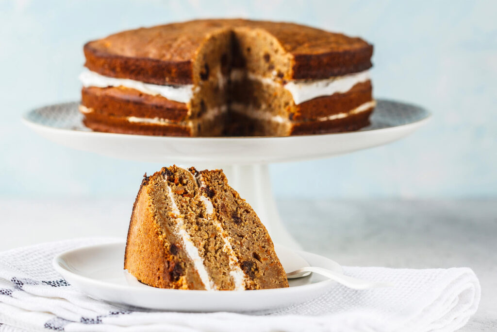 Veganer Karottenkuchen mit Kokoscreme auf einer Tortenplatte, davor ein Stück mit drei Böden und zwei Cremeschichten