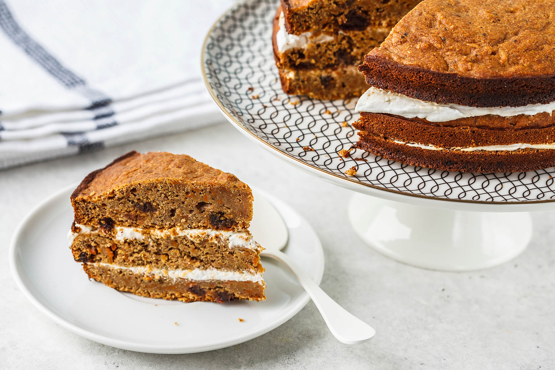 Ein Stück veganer Karottenkuchen mit Kokoscreme auf einem Teller, dahinter die angeschnittene Torte