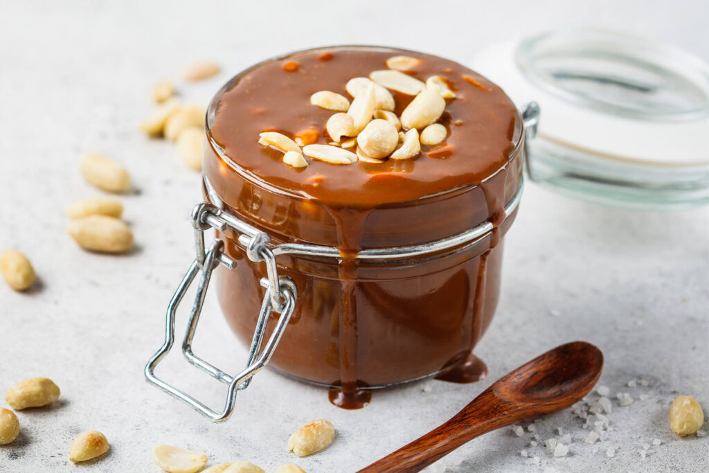 Dickes veganes Kokos-Karamell im Glas mit hellen Erdnusskernen, Salzflocken und Holzlöffel daneben