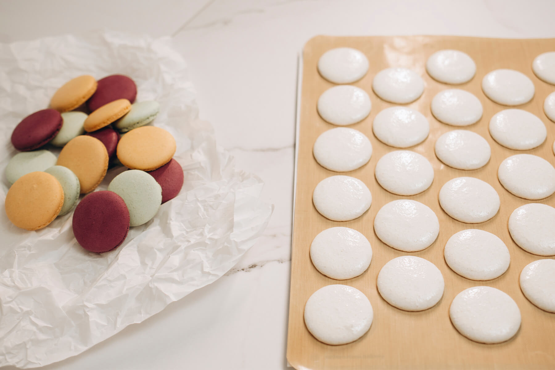 Eine Backmatte mit dicht an dicht gespritzten hellen Macaronschalen in gleichmäßigen Reihen, daneben liegen fertige bunte Macarons auf Seidenpapier