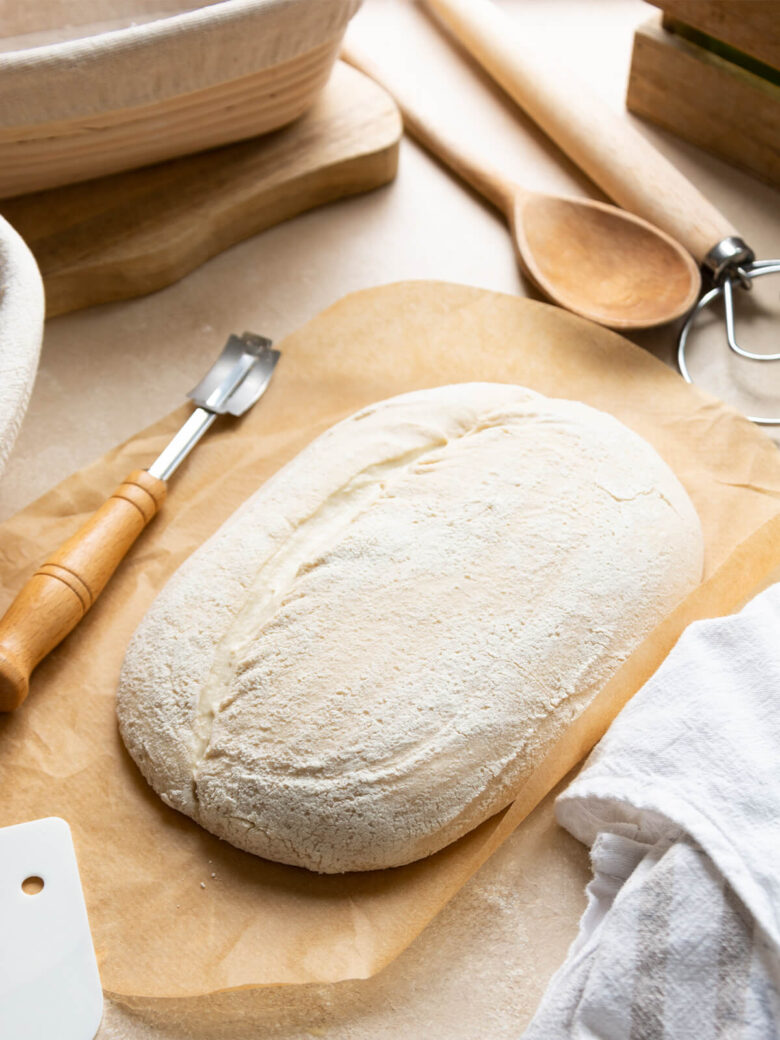 Ein Bäckermesser mit Holzgriff liegt auf Backpapier neben einem bemehlten ovalen Brotteigling mit einem langen Schnitt, dahinter Gärkörbe, ein Holzbrett, ein Holzlöffel und ein Teigrührer, vorn eine Teigkarte