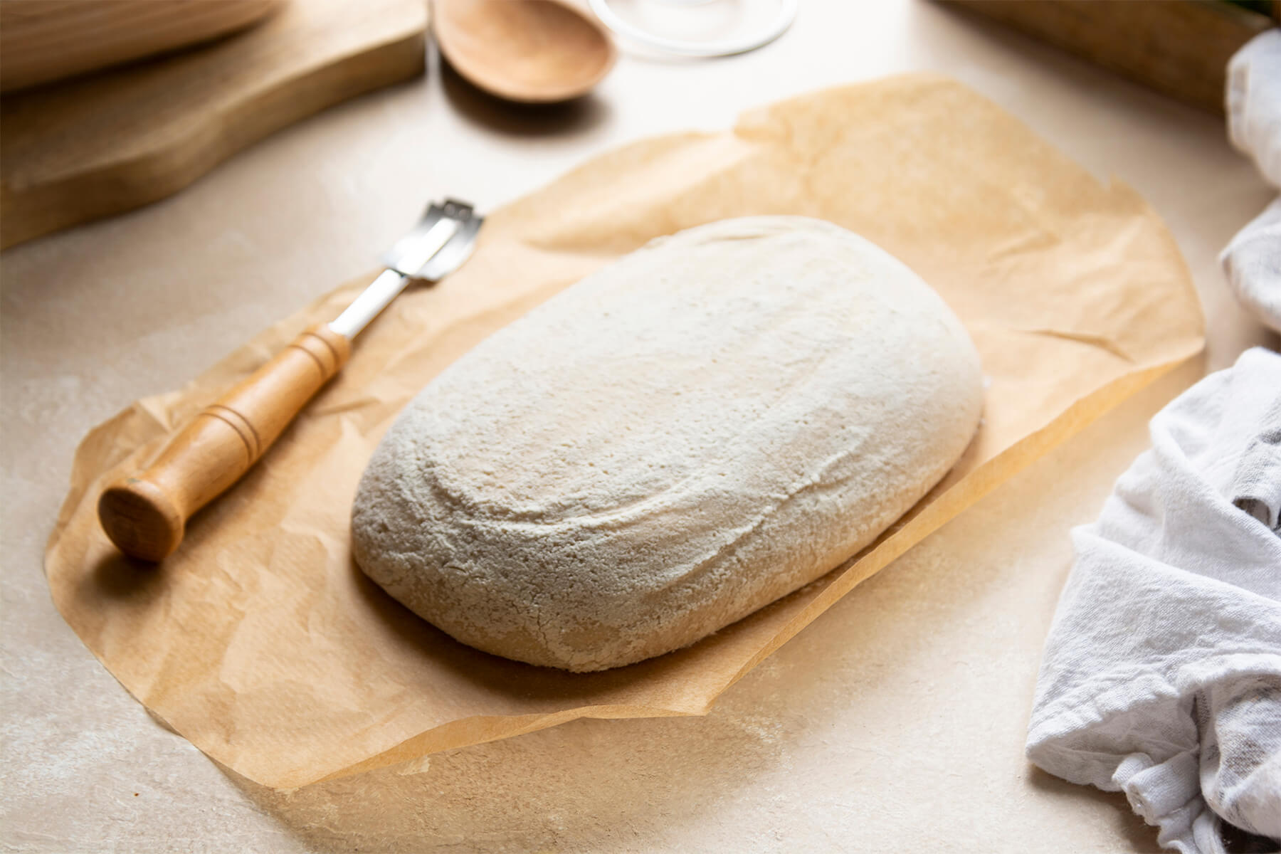 Ein Bäckermesser mit gedrechseltem Holzgriff und gebogener Klinge liegt neben einem stark bemehlten, länglichen Brotteigling auf Backpapier