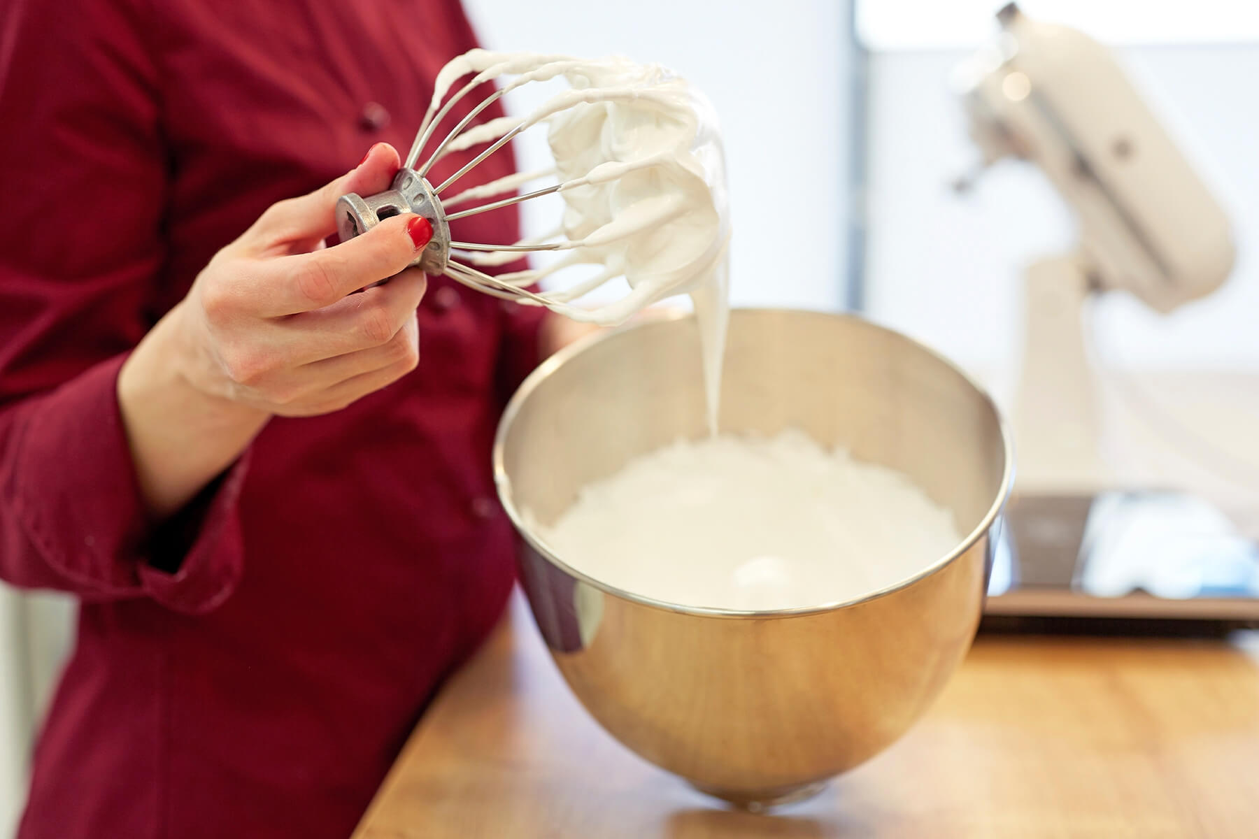 Ein abgenommener Schneebesen wird über einer Schüssel gehalten, an den Drähten hängt weiße glänzende Baisermasse und bildet weiche Spitzen