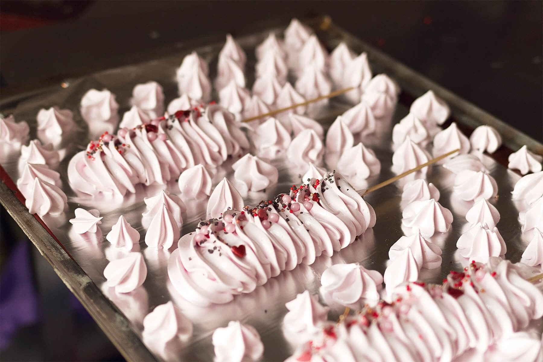 Backblech mit Alufolie, darauf mehrere rosa Baiserraupen mit Streudeko und viele kleine spitze rosa Baisertupfen dazwischen, ein Tupfen liegt umgekippt auf der Seite