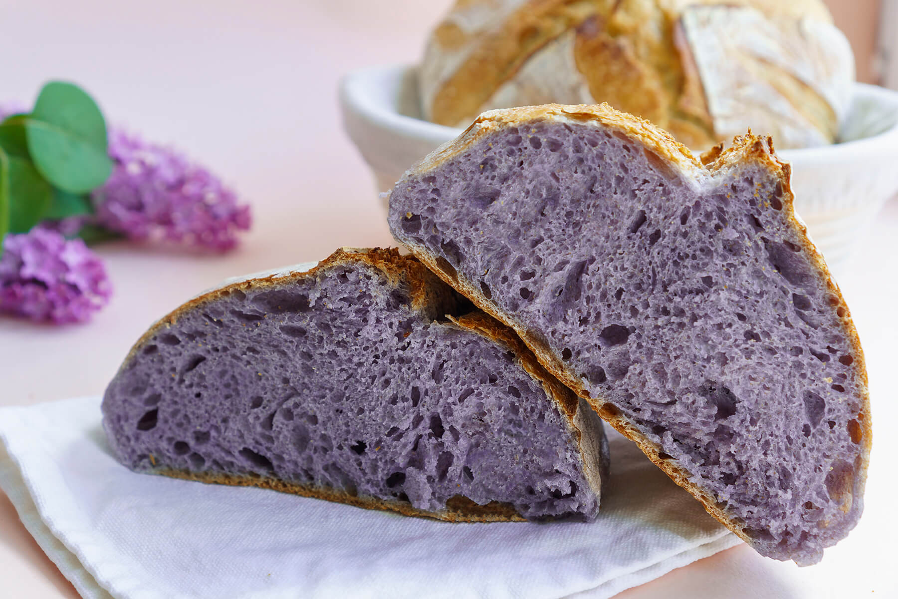 Zwei Hälften eines angeschnittenen runden Brotes mit durchgehend lila-violetter, offenporiger Krume und dünner goldbrauner Kruste auf einem weißen Tuch, daneben Fliederblüten