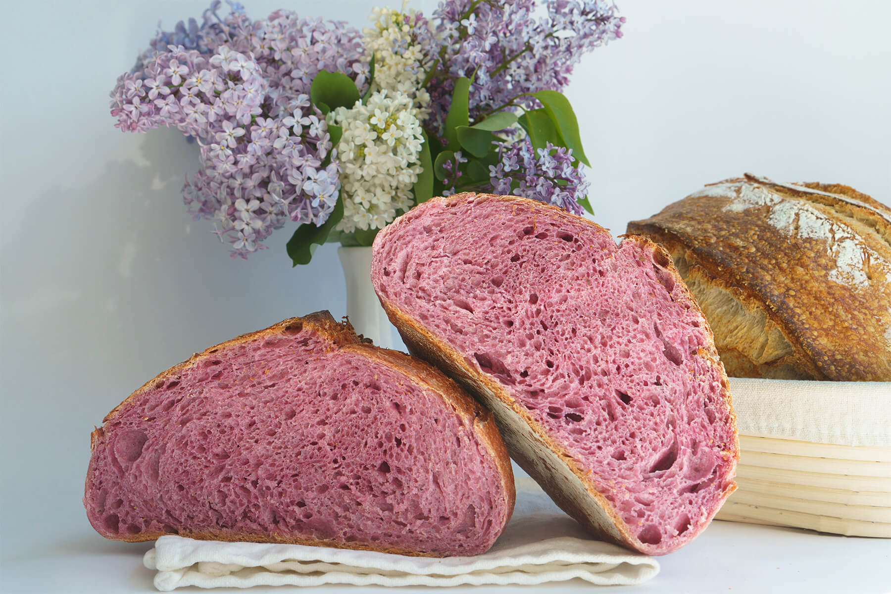 Zwei Hälften eines angeschnittenen Brotes mit kräftig rosa gefärbter Krume vor einem Fliederstrauß, dahinter ein helles Brot auf einem Gärkörbchen