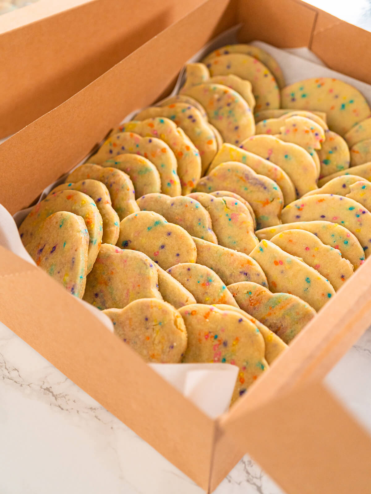 Bunte Plätzchen mit Zuckerstreuseln im Teig, in dichten Reihen schräg aneinandergelehnt, in einem mit Backpapier ausgelegten Karton aus braunem Kraftpapier