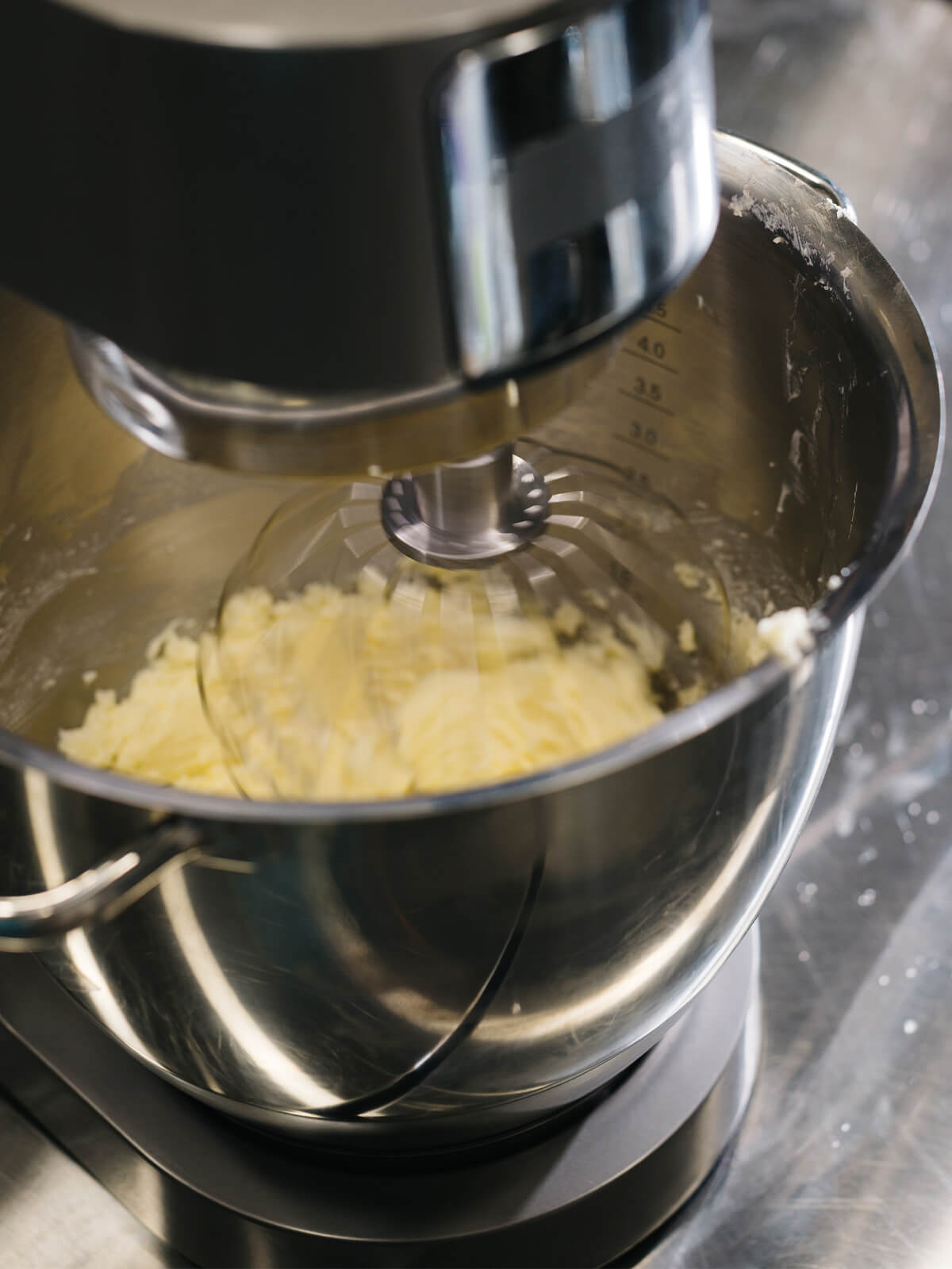 In der Rührschüssel einer Küchenmaschine liegt eine helle, deutlich körnig-flockige Buttermasse am Schneebesen
