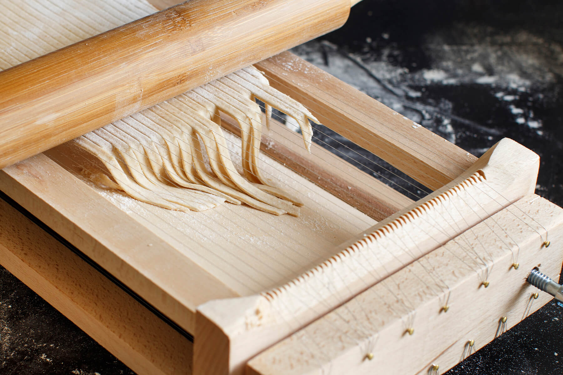 Holzrahmen einer Chitarra mit gespannten Stahlsaiten, gekerbter Halteleiste und seitlicher Spannschraube, darauf frisch geschnittene Teigstränge