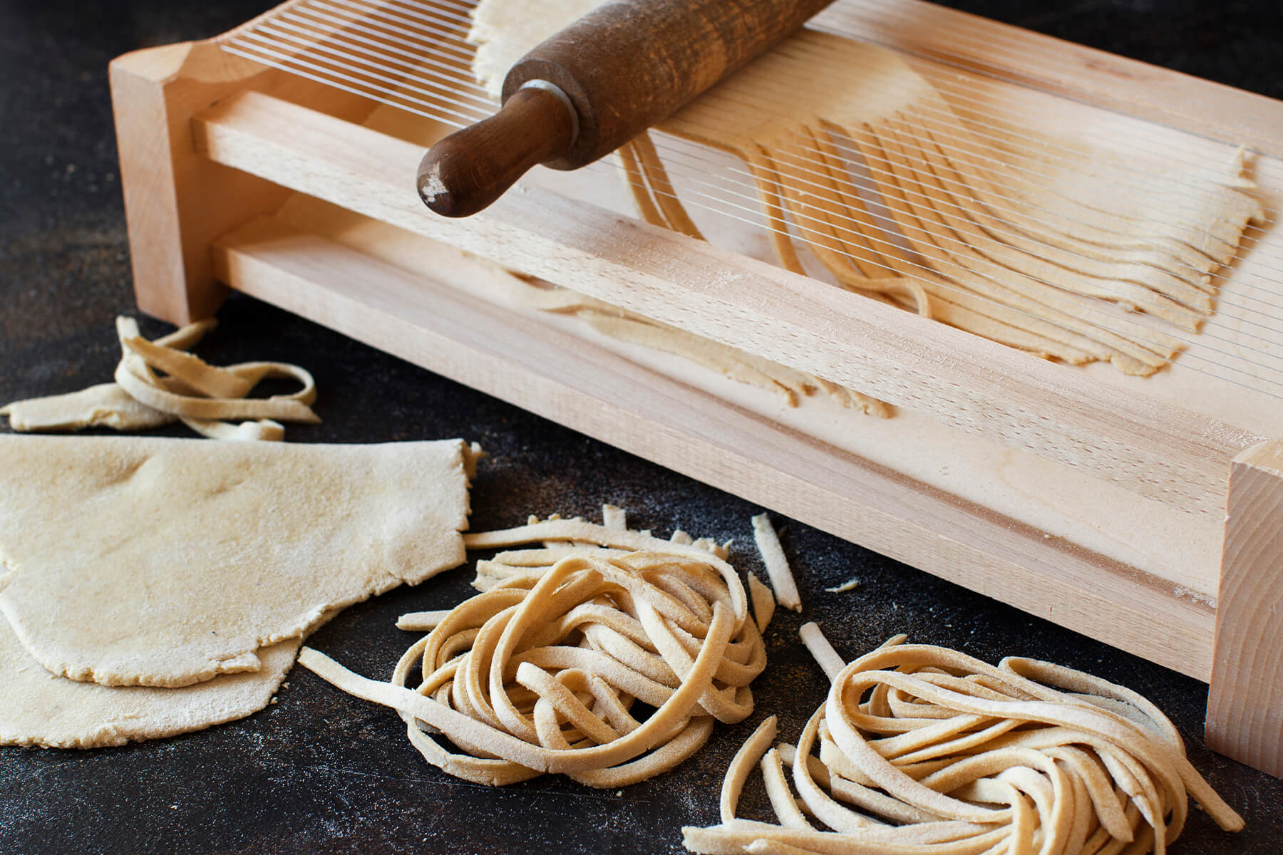Hölzerner Chitarra-Rahmen mit zwei unterschiedlich dicht bespannten Saitenfeldern, darauf eine Teigbahn unter einem Nudelholz, davor zwei Nester fertiger Stränge