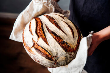 Zwei Hände halten in einem Leinentuch ein rundes, bemehltes Sauerteigbrot mit langen, gebogenen Schnitten, die sich im Ofen weit geöffnet haben und tief rotbraun bis fast schwarz ausgebacken sind