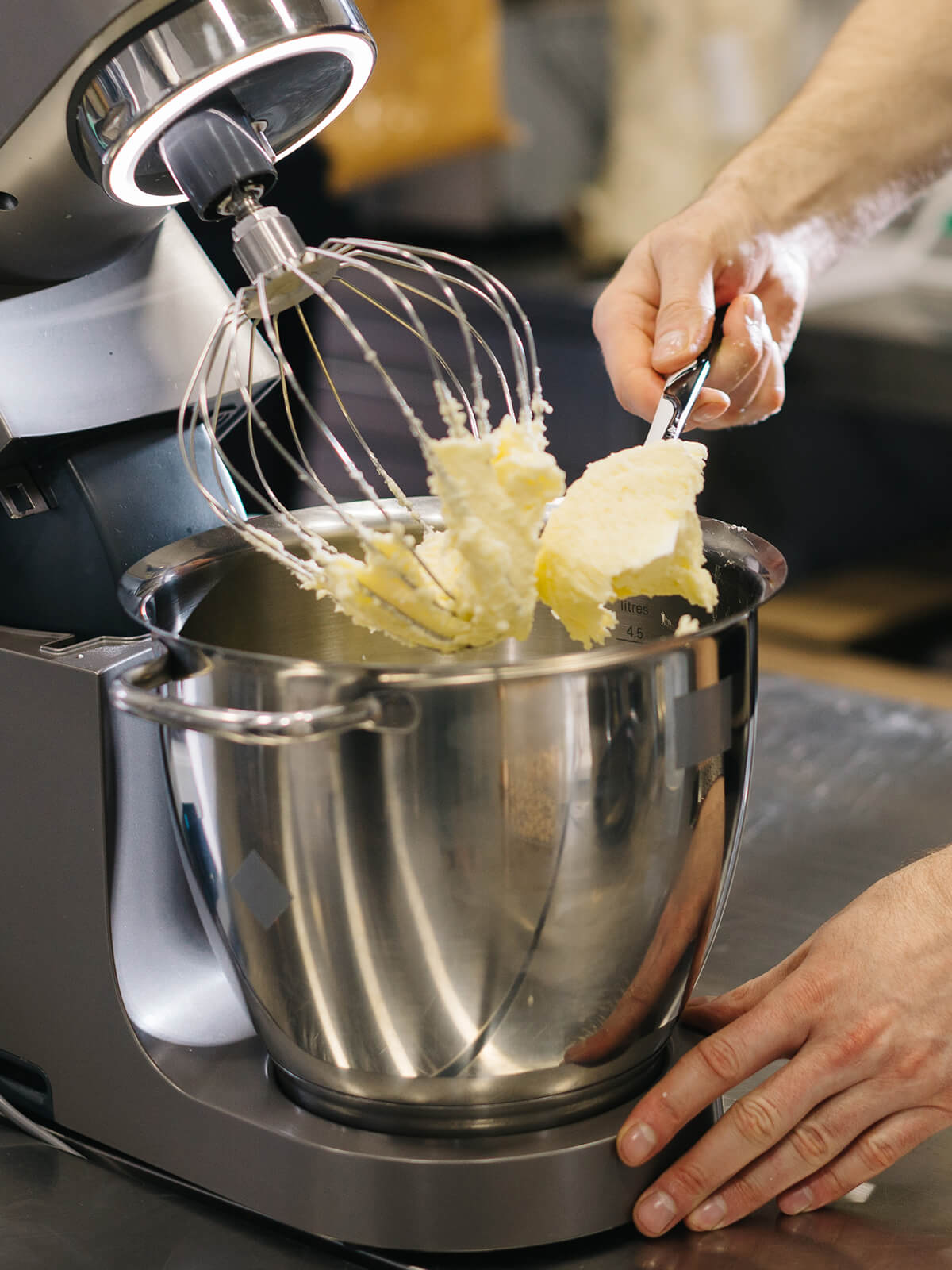 Am abgehobenen Schneebesen einer Küchenmaschine hängt eine helle, dichte Buttercreme, eine Hand streift sie mit einem Spatel ab