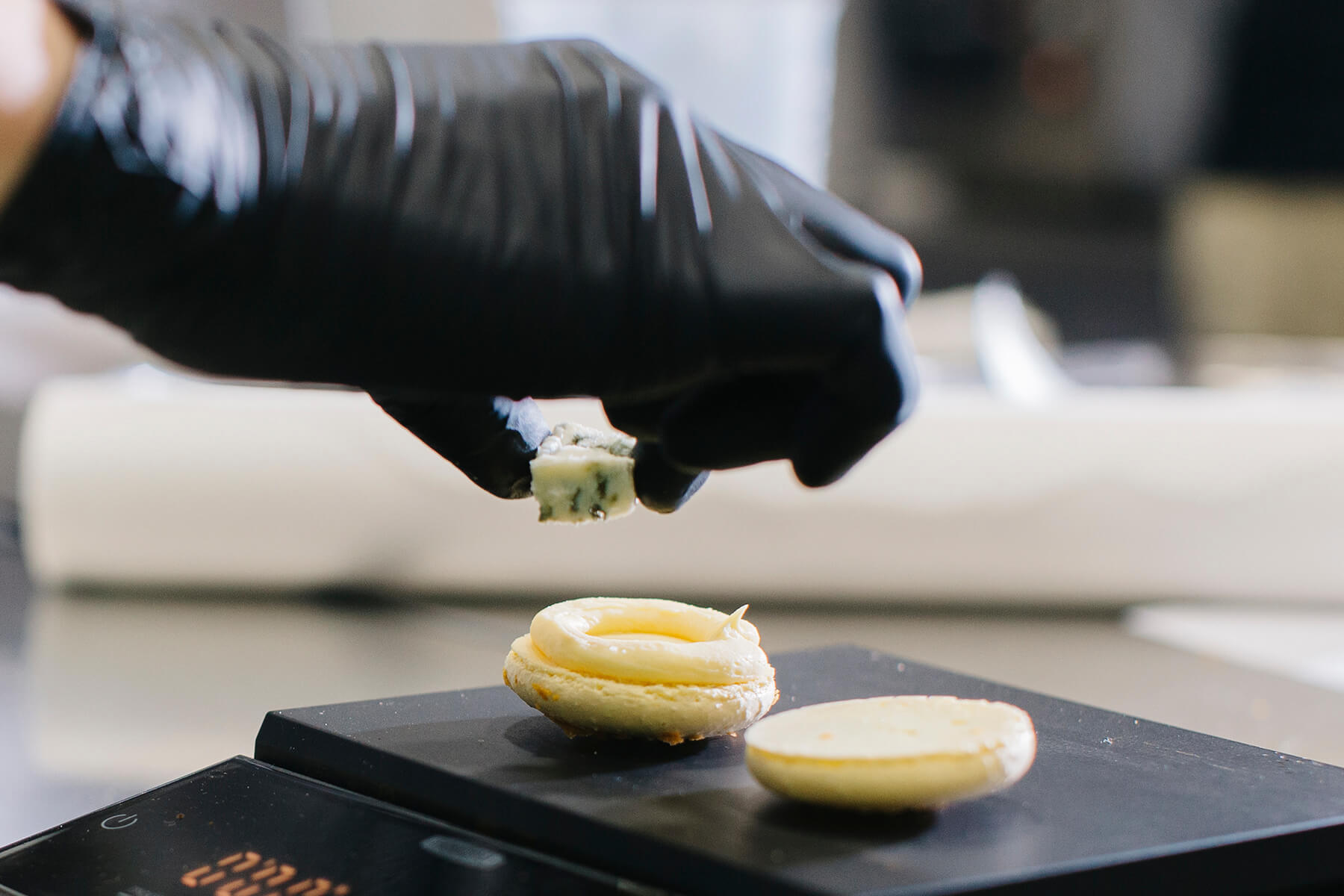 Auf einer digitalen Waage liegen eine Macaronschale mit aufgespritzter Creme und eine leere Schale, eine behandschuhte Hand hält etwas darüber