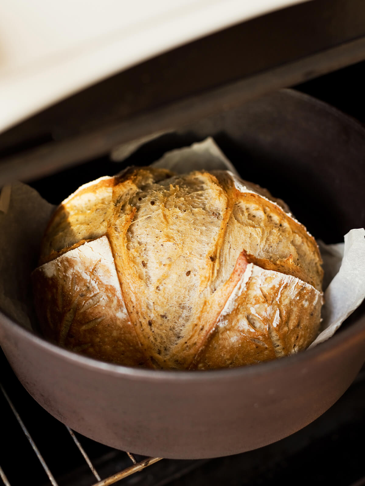 Frisch gebackenes, eingeschnittenes Sauerteigbrot auf Backpapier in einem dunklen, schweren Topf im Backofen, darüber der angehobene Deckel
