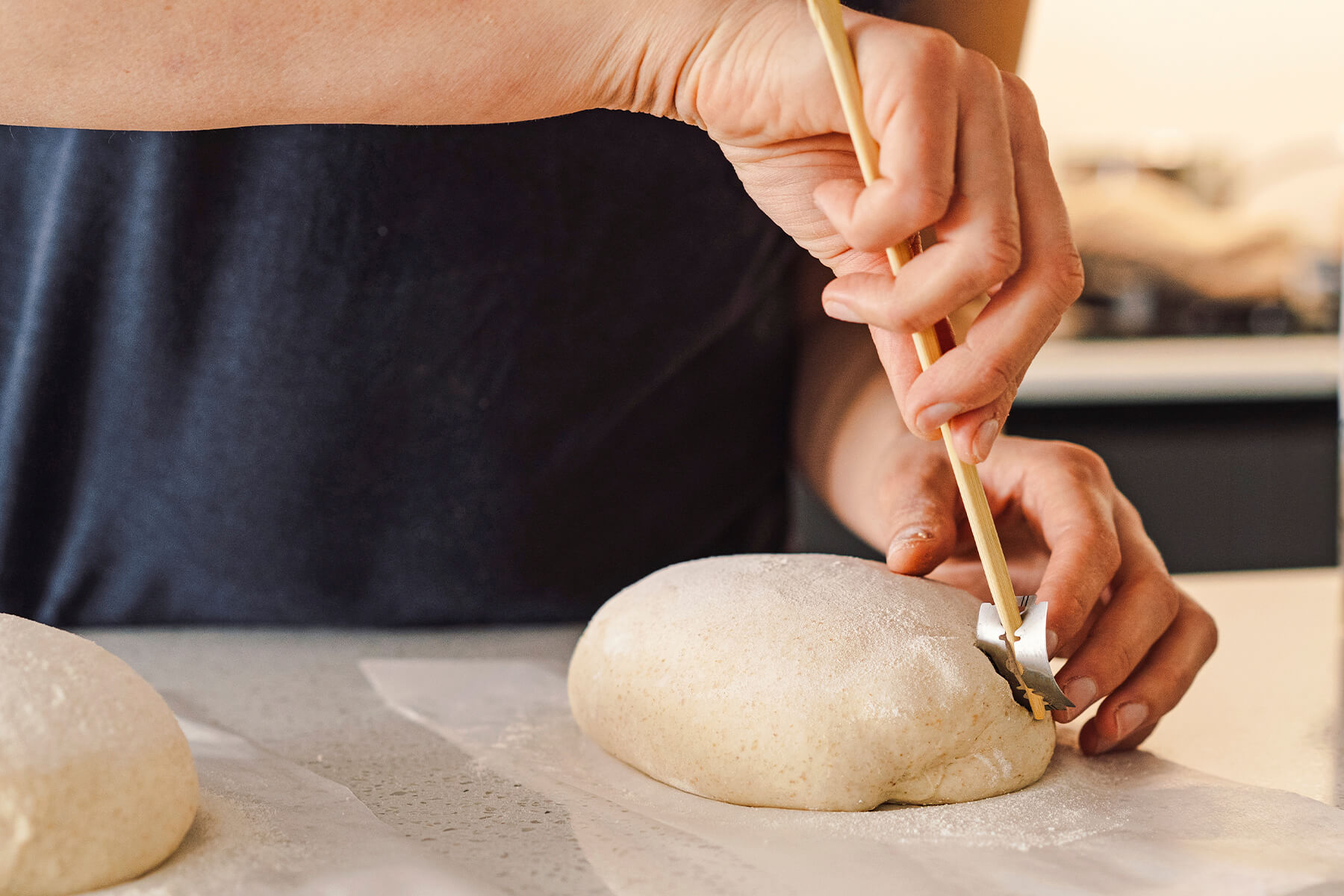 Eine Hand führt eine gebogene Klinge auf einem Holzstab an einen bemehlten länglichen Teigling heran und setzt den ersten Schnitt