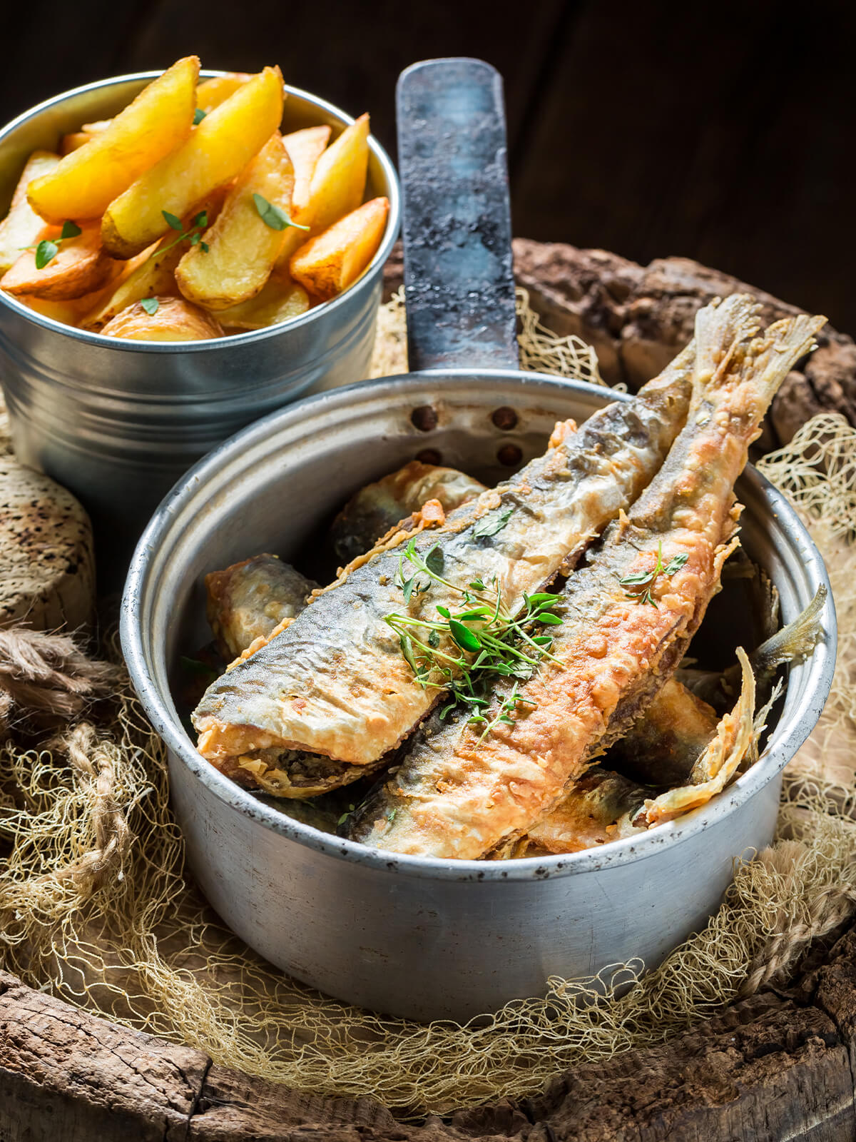 Ausgebackene Sardinen mit knuspriger Kruste und frischem Thymian in einem kleinen Metalltopf, dahinter goldbraune Kartoffelspalten in einem Metallbecher