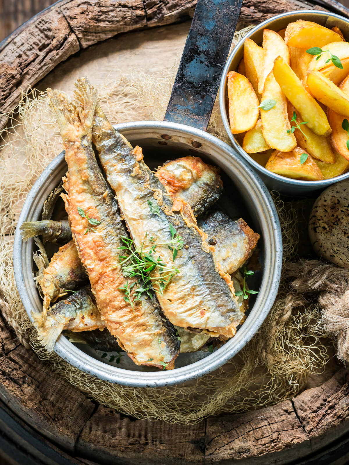 Nahaufnahme knusprig ausgebackener Sardinen mit goldbrauner Kruste und Thymian, daneben Kartoffelspalten