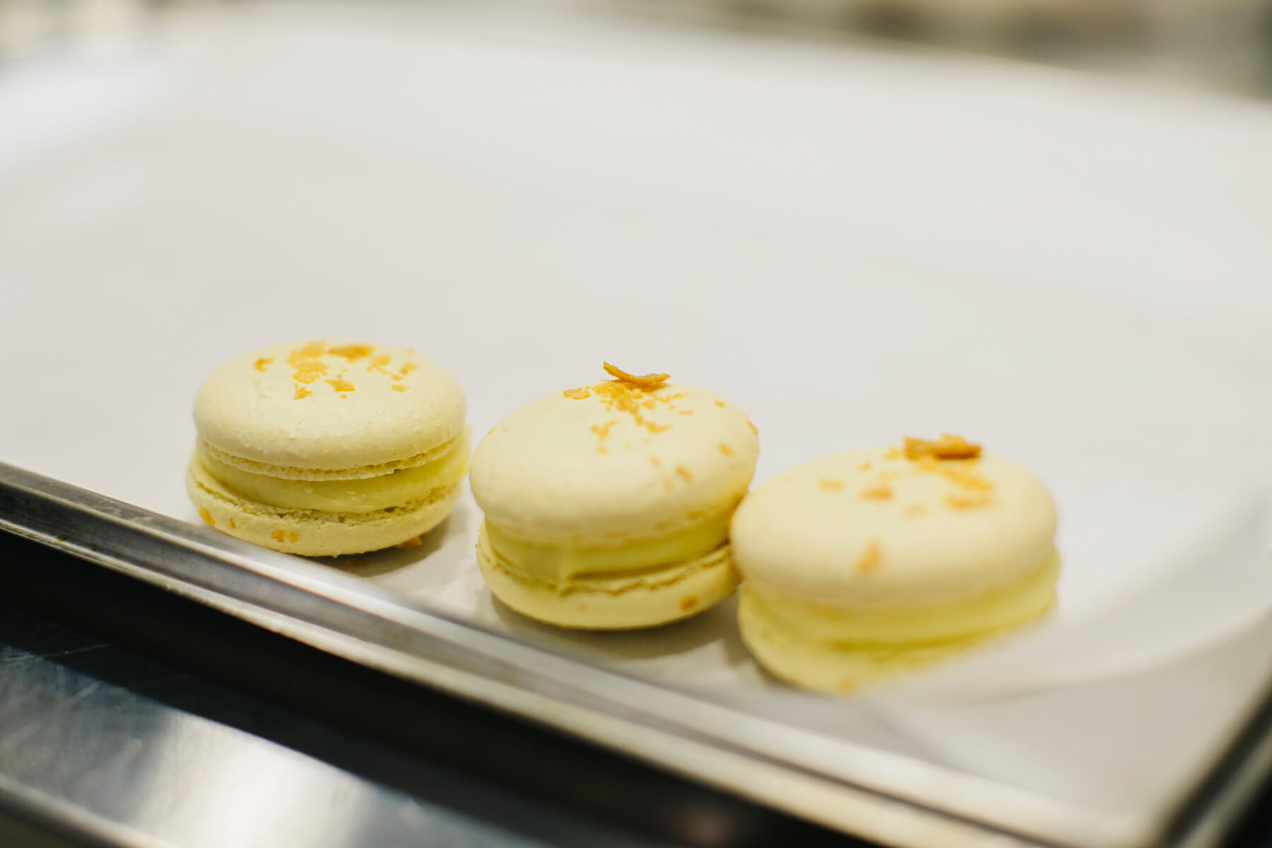 Drei fertige Macarons stehen nebeneinander auf einem Blech, jeder mit glatter Haube, gekräuseltem Fuß und einer sichtbaren Cremeschicht dazwischen