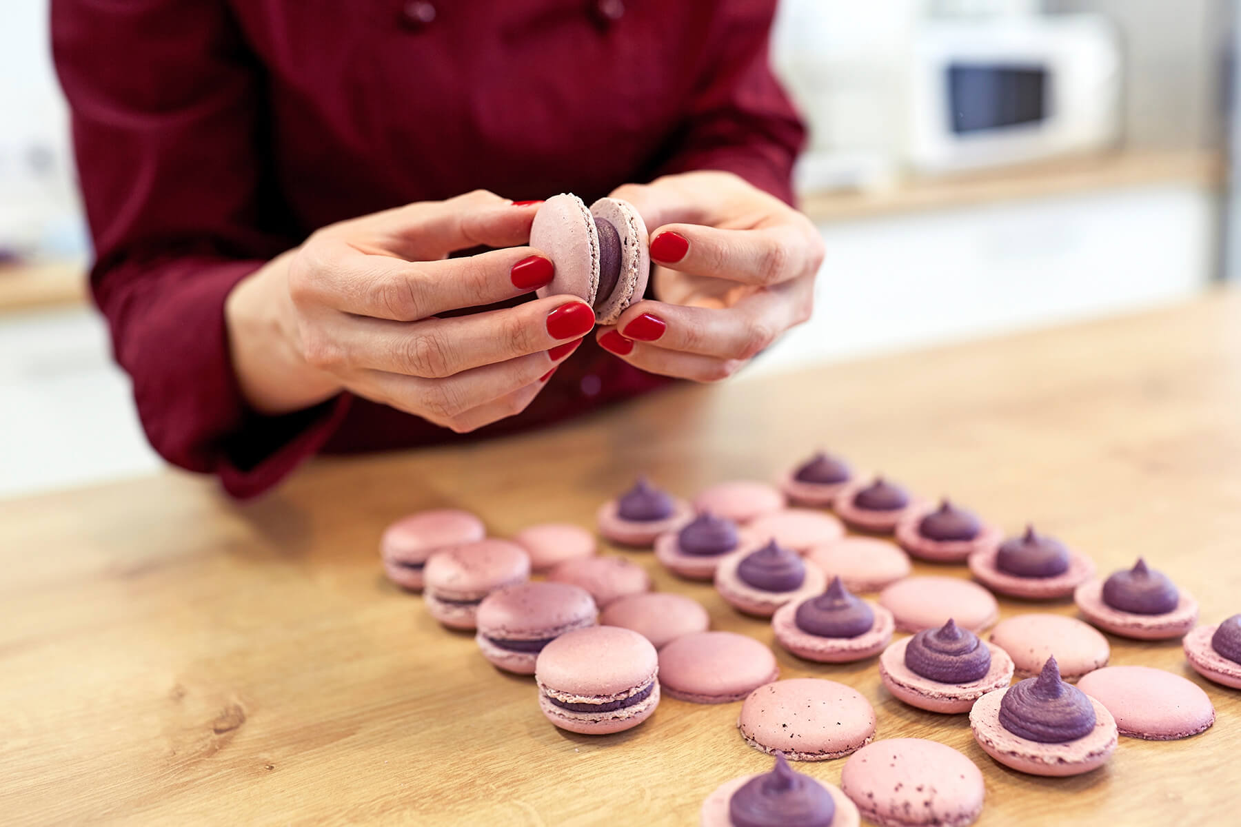 Zwei Hände halten ein fertig zusammengesetztes rosafarbenes Macaron, davor liegen Schalen mit violetten Cremetupfen in Reihen