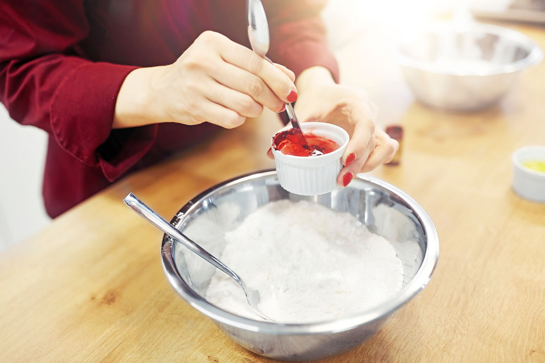 Ein Löffel nimmt rote Gelfarbe aus einer kleinen Schale, daneben steht eine Schüssel mit weißem Puderzucker und einem Löffel