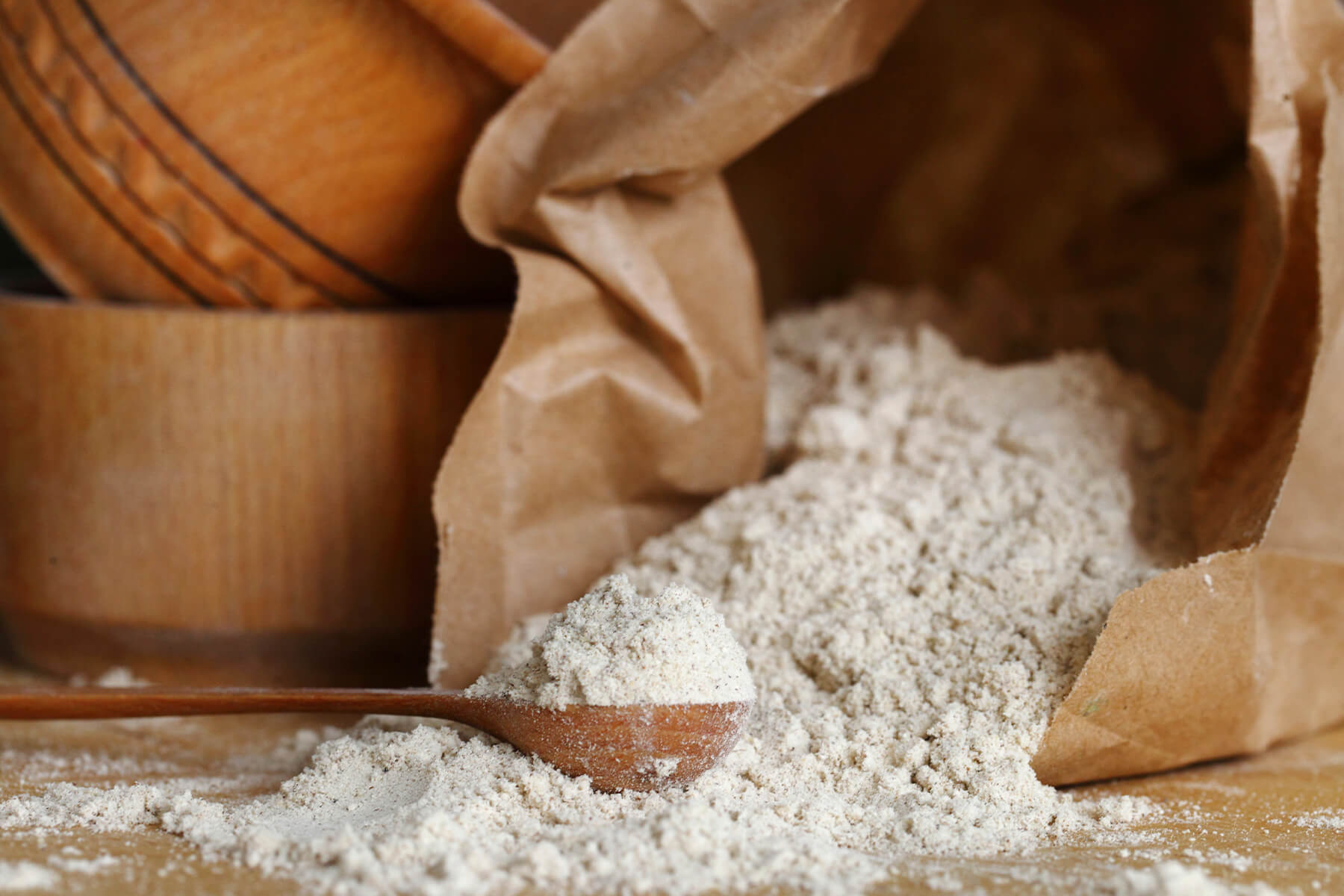 Umgekippte braune Papiertüte auf einem Holztisch, daraus ausgelaufenes grau-beiges Mandelmehl mit feinen dunklen Pünktchen, davor ein Holzlöffel mit einer gehäuften Portion, links im Anschnitt zwei Holzschalen