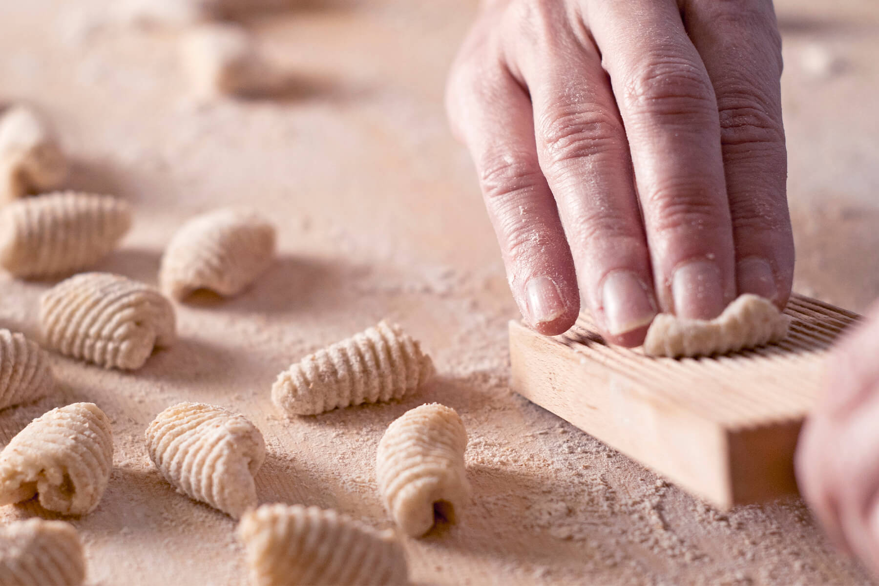 Hand rollt ein Teigstück über ein leicht bemehltes Gnocchi-Brett, ringsum liegen fertige Stücke