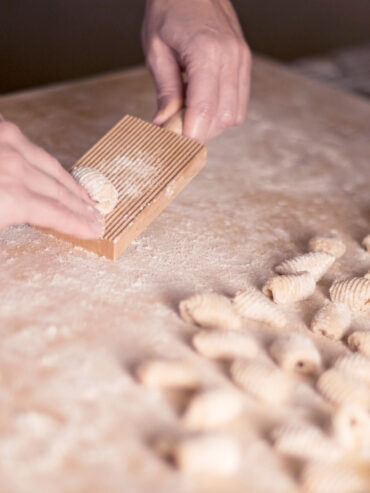 Hand rollt ein Teigstück über ein geriffeltes Gnocchi-Brett, daneben liegen fertig geformte Stücke