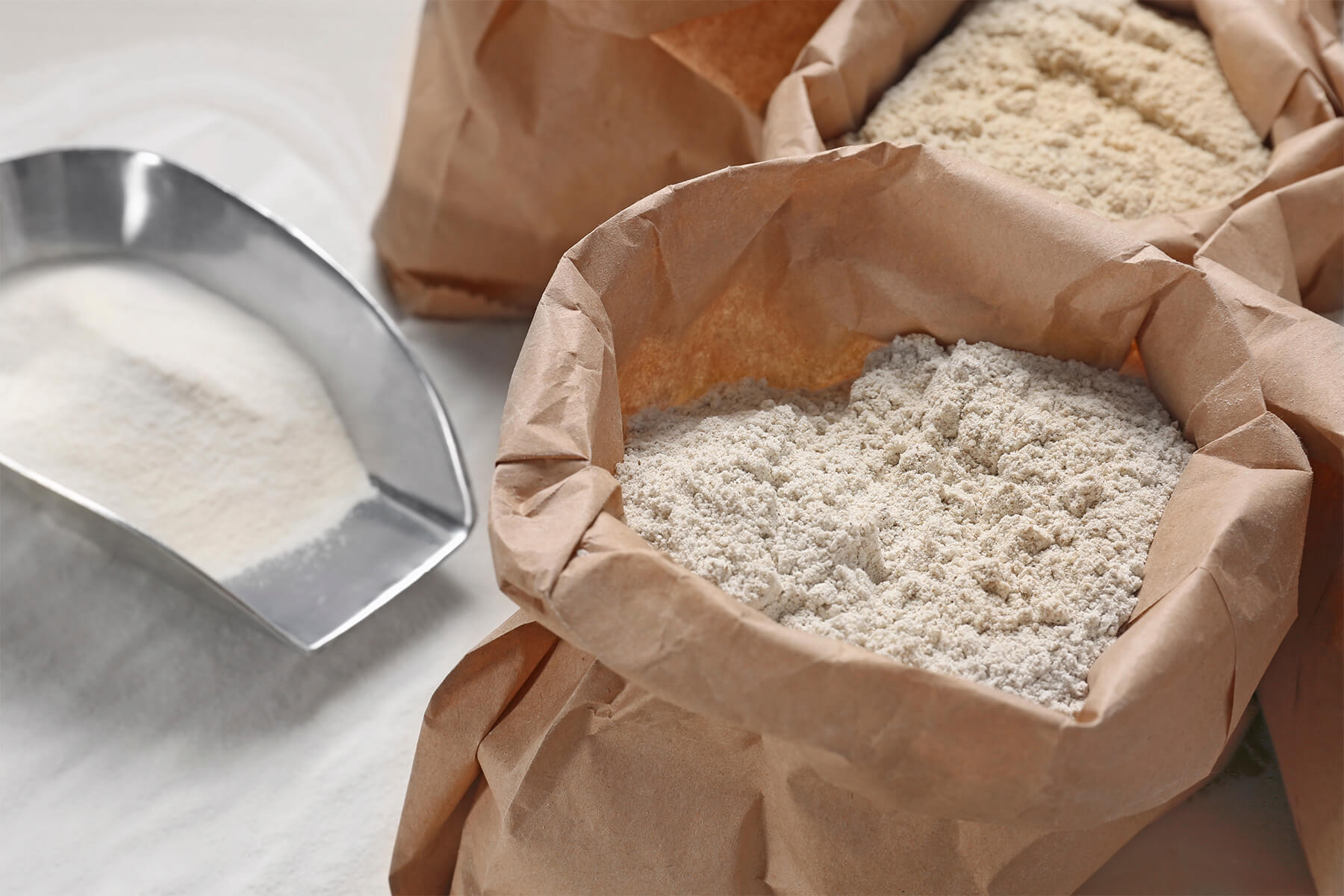 Metallschaufel mit schneeweißem Mehl, daneben ein offener Papiersack mit graubeigem, fein gesprenkeltem Mehl, dahinter ein Sack mit beigem Mehl