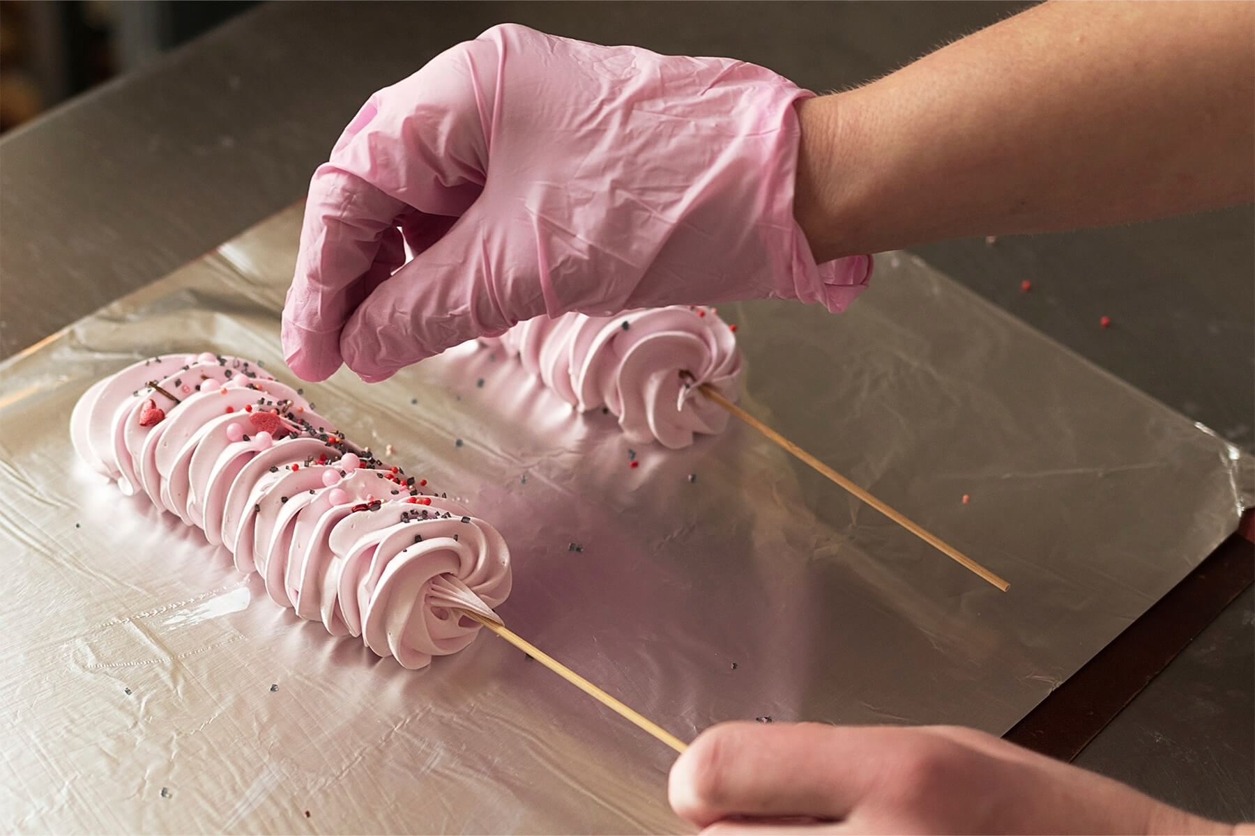 Eine Hand in rosa Handschuh streut Zuckerperlen und Streusel auf eine frisch gespritzte rosa Baiserraupe, in deren Rosette am Ende ein Holzspieß steckt, die andere Hand hält den Spieß
