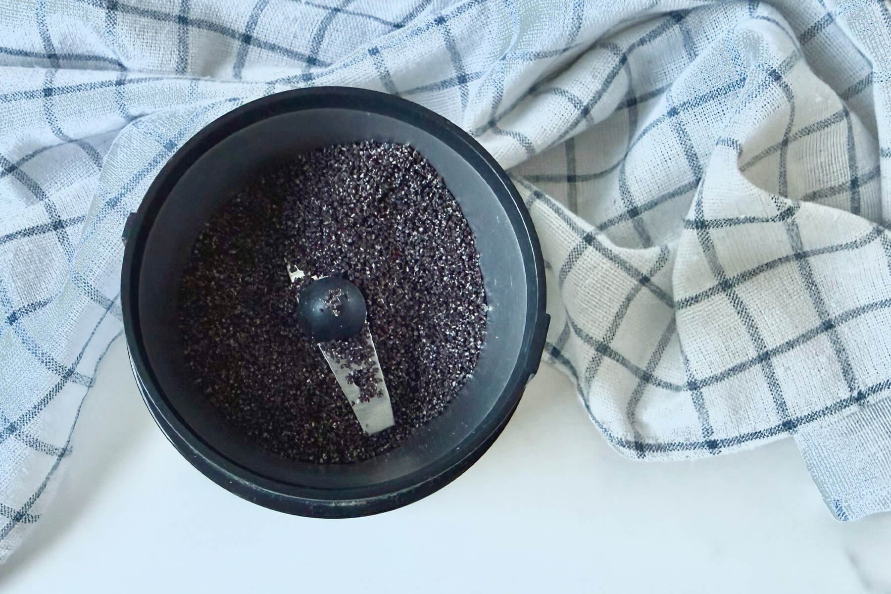 Blick von oben in eine geöffnete Kaffeemühle mit Schlagmesser, darin gemahlener Blaumohn mit aufgebrochenen Körnern, ringsum ein blau-weiß kariertes Küchentuch