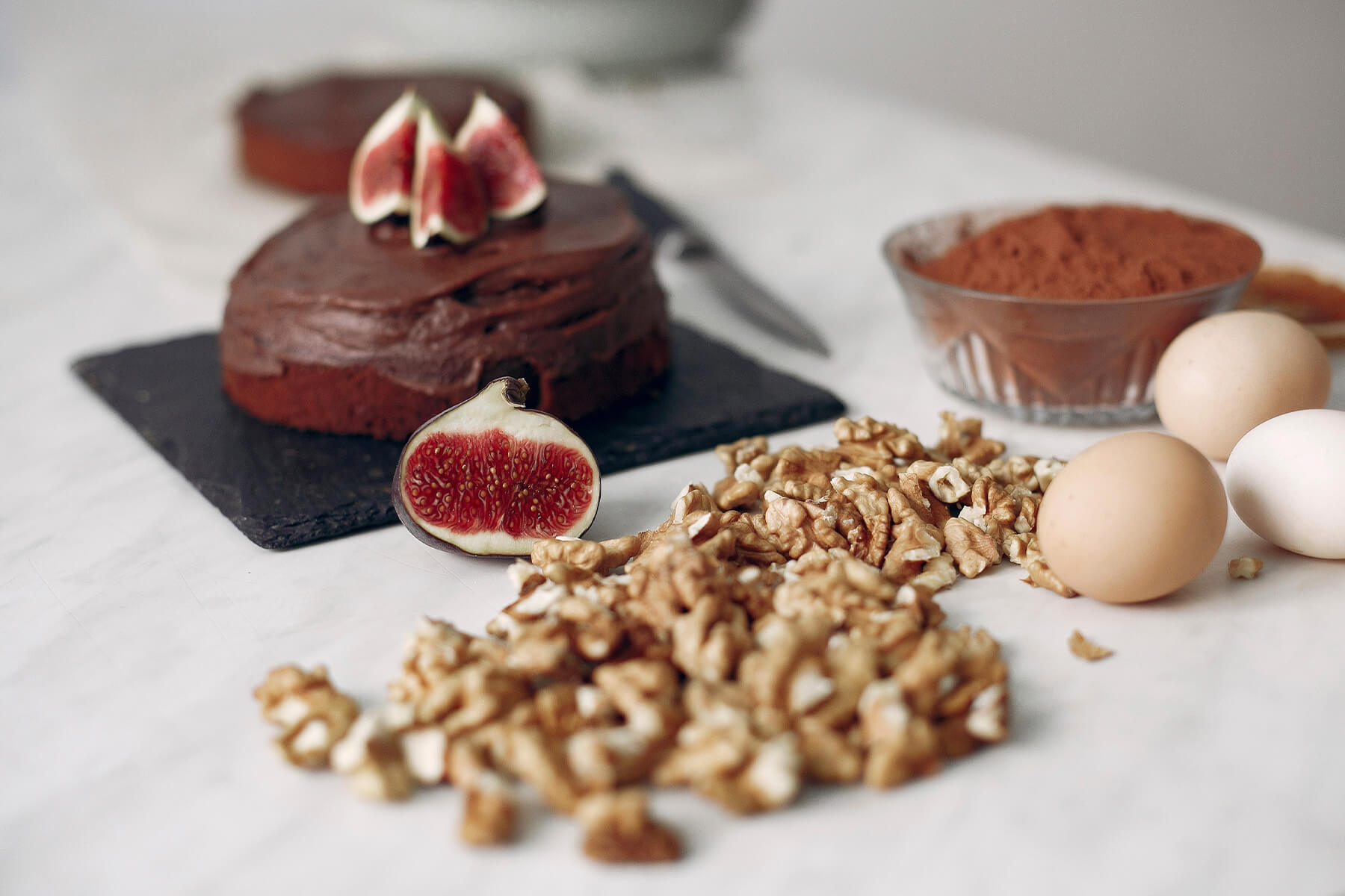 Eine Glasschale mit Kakaopulver neben braunen Eiern, Walnusskernen und einem Schokoladenkuchen auf einer Schieferplatte