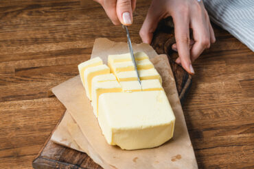 Hände schneiden ein festes Stück Butter auf Backpapier und einem dunklen Holzbrett mit dem Messer in Scheiben und Würfel
