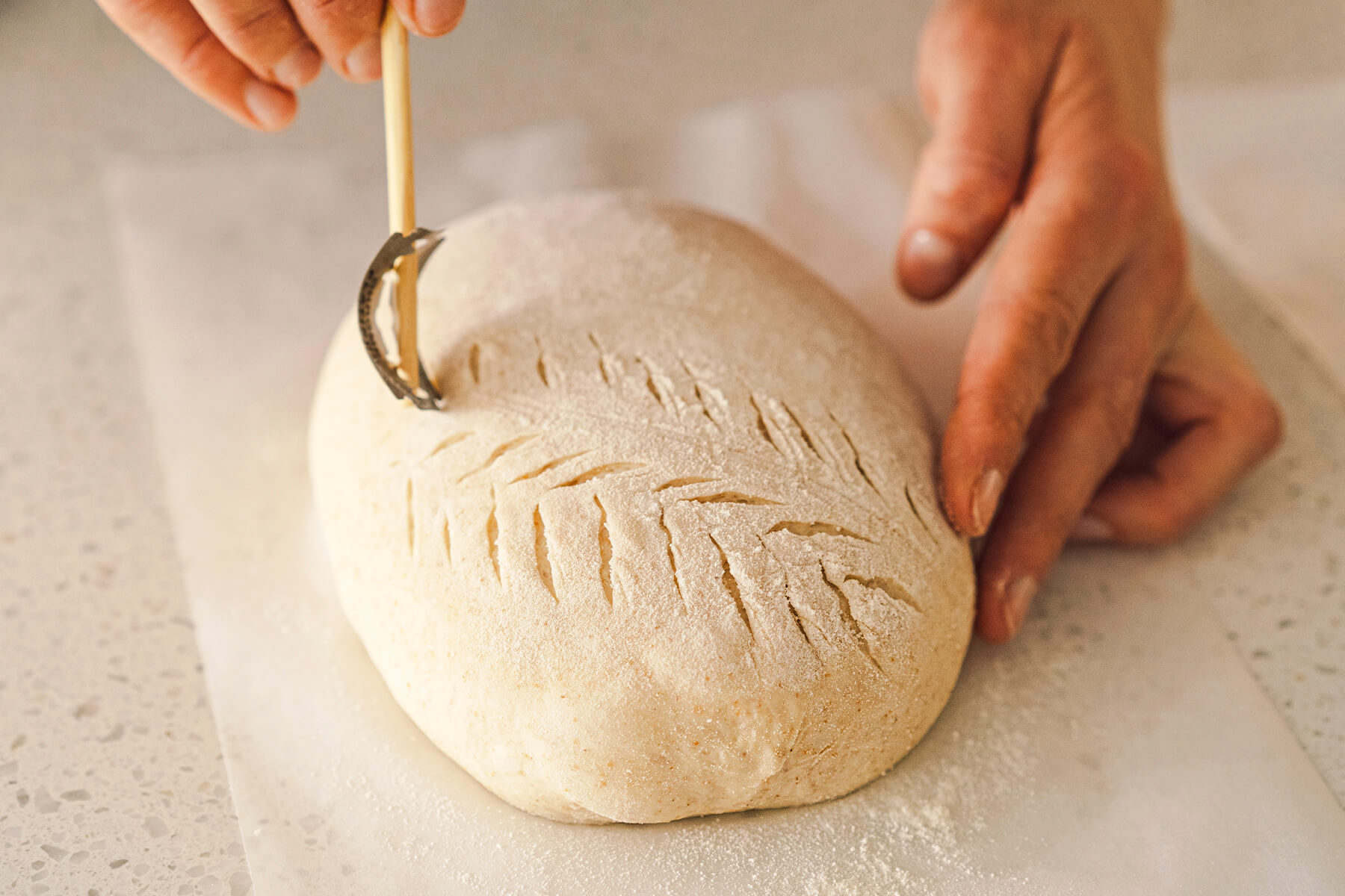Nahaufnahme eines Ährenmusters auf bemehltem Teig, die gebogene Klinge am Holzstab setzt eine einzelne schräge Kerbe neben der Mittellinie