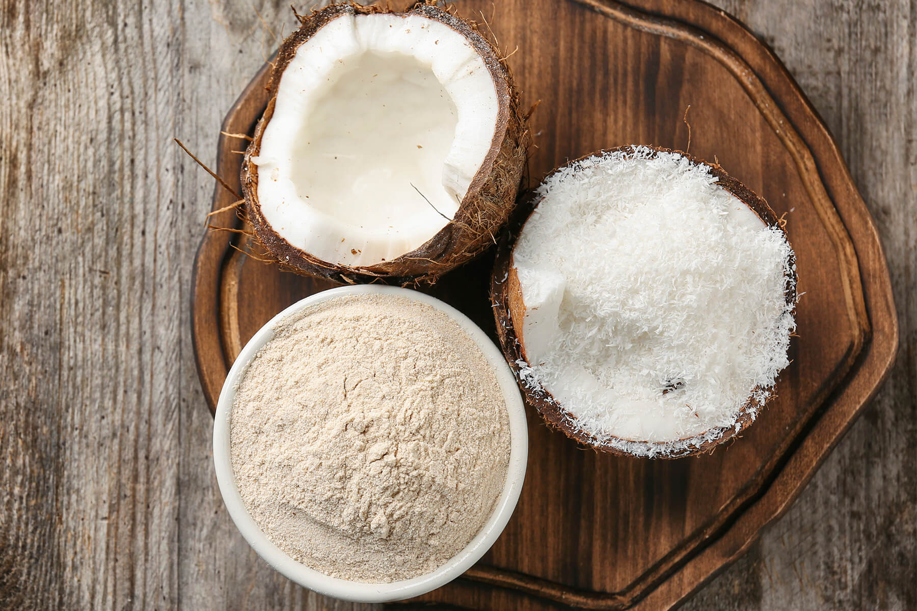 Weiße Schale mit hellbeigem Kokosmehl auf einem dunklen Holzbrett, daneben eine leere Kokosnusshälfte und eine zweite, gefüllt mit feinen weißen Kokosraspeln