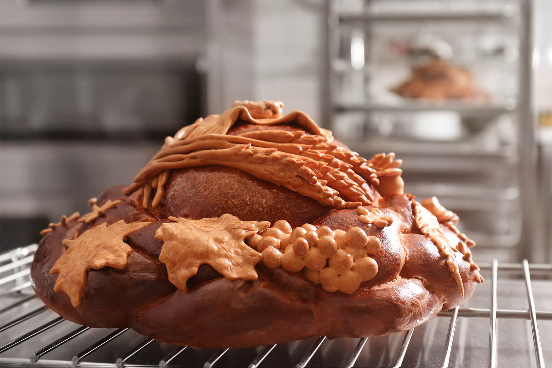 Gebackenes Hochzeitsbrot auf einem Gitter in der Backstube, heller Teigschmuck mit Ährengarbe, Tuch, Traube und Blättern auf dem tief rotbraunen, glänzenden Laib, im Hintergrund ein Regal mit einem weiteren Brot