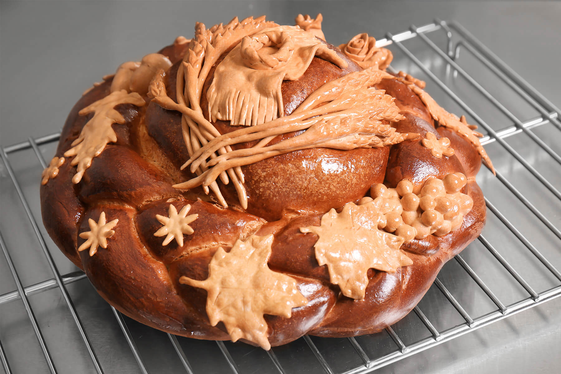 Gebackenes Hochzeitsbrot auf einem Kuchengitter: tief rotbraun glänzender Zopfkranz mit Kuppel, darauf heller Schmuck aus Teig mit Ährengarben, einem Tuch mit Fransen, Rosen, einer Traube, Blättern und Sternen