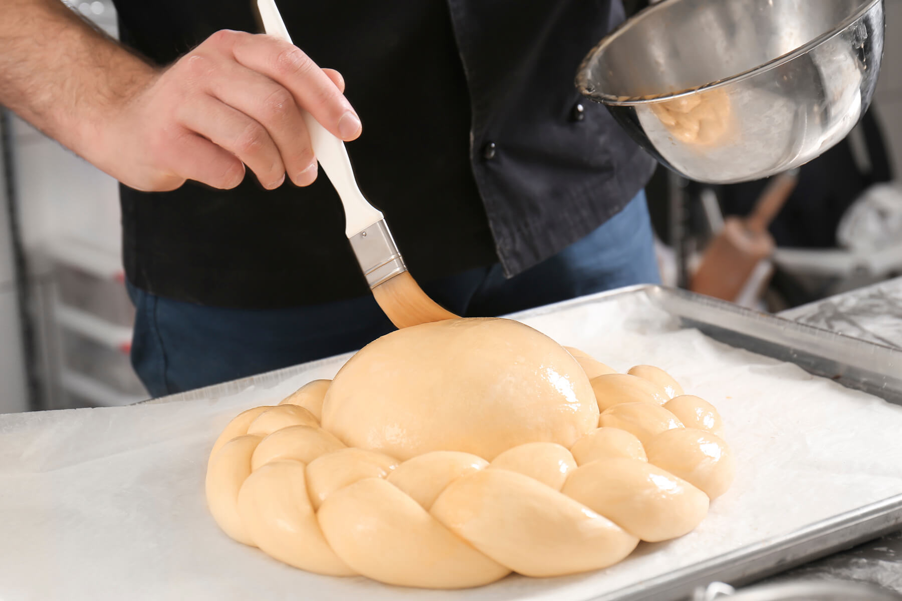 Eine Hand bestreicht die rohe Teigkuppel eines Hochzeitsbrots mit dem Pinsel, ringsum liegt ein geflochtener Kranz aus Hefeteig auf einem Blech mit Backpapier, die andere Hand hält eine Schüssel mit Eistreiche