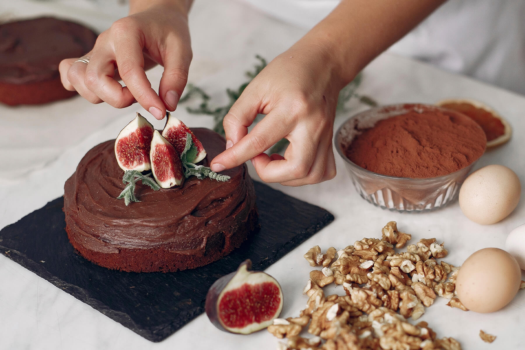 Zwei Hände setzen Feigenviertel auf einen mit Schokoladencreme bestrichenen Kuchen, daneben Kakaopulver in einer Glasschale und Walnusskerne