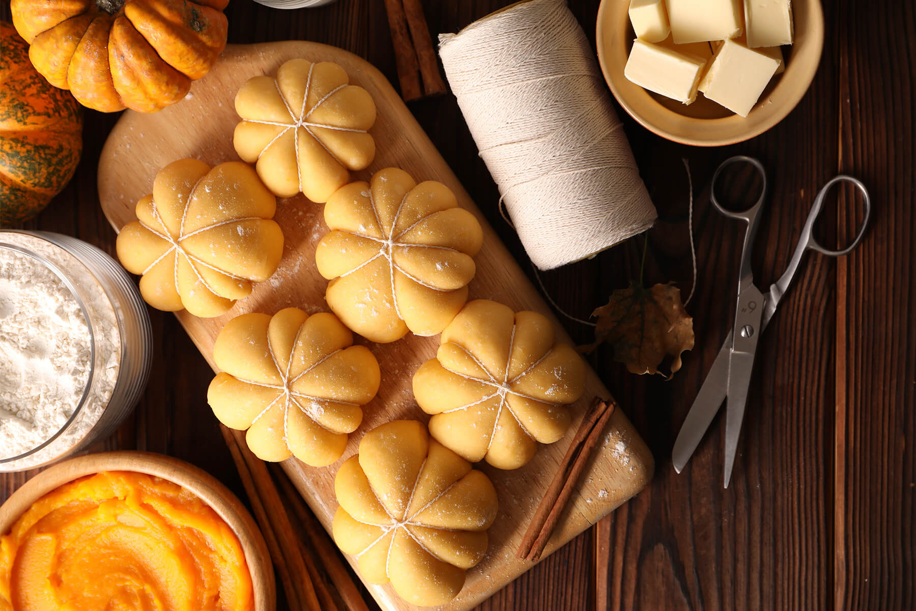 Sechs rohe, mit weißem Küchengarn gebundene Hefebrötchen auf einem Holzbrett, daneben eine Rolle Küchengarn, eine Schere, Butterwürfel, eine Schale Kürbispüree und ein Glas Mehl