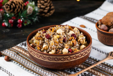 Geschnitzte Holzschale mit Kutia aus Weizenkörnern, roten Cranberrys, ganzen Haselnüssen, Walnussstücken und Mohnkörnern, daneben Tannenzapfen, eine Schale mit Nüssen und weihnachtlicher Tischschmuck