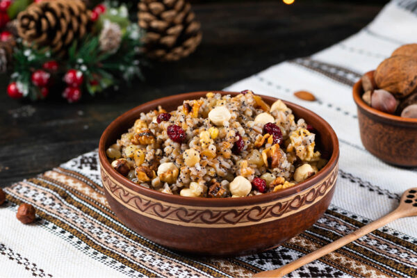 Geschnitzte Holzschale mit Kutia aus Weizenkörnern, roten Cranberrys, ganzen Haselnüssen, Walnussstücken und Mohnkörnern, daneben Tannenzapfen, eine Schale mit Nüssen und weihnachtlicher Tischschmuck