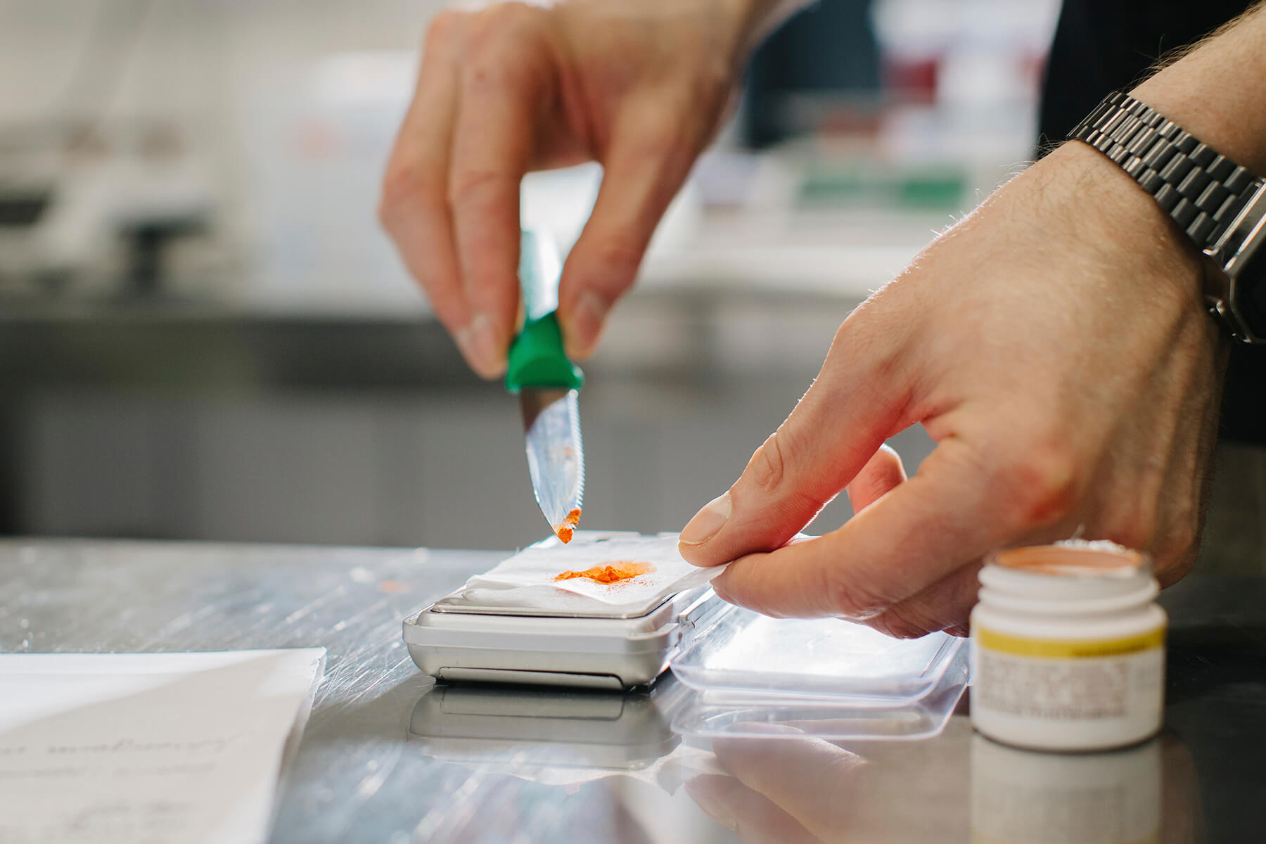 Mit einem kleinen grünen Spatel wird orangefarbene Lebensmittelfarbe auf eine Feinwaage aufgetragen, daneben steht ein Farbtöpfchen