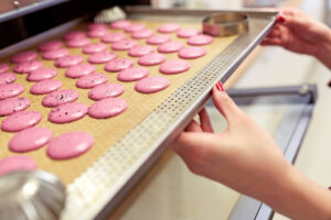 Zwei Hände schieben ein Lochblech mit einer Backmatte und vielen rosafarbenen Macaronschalen in versetzten Reihen in einen Ofen