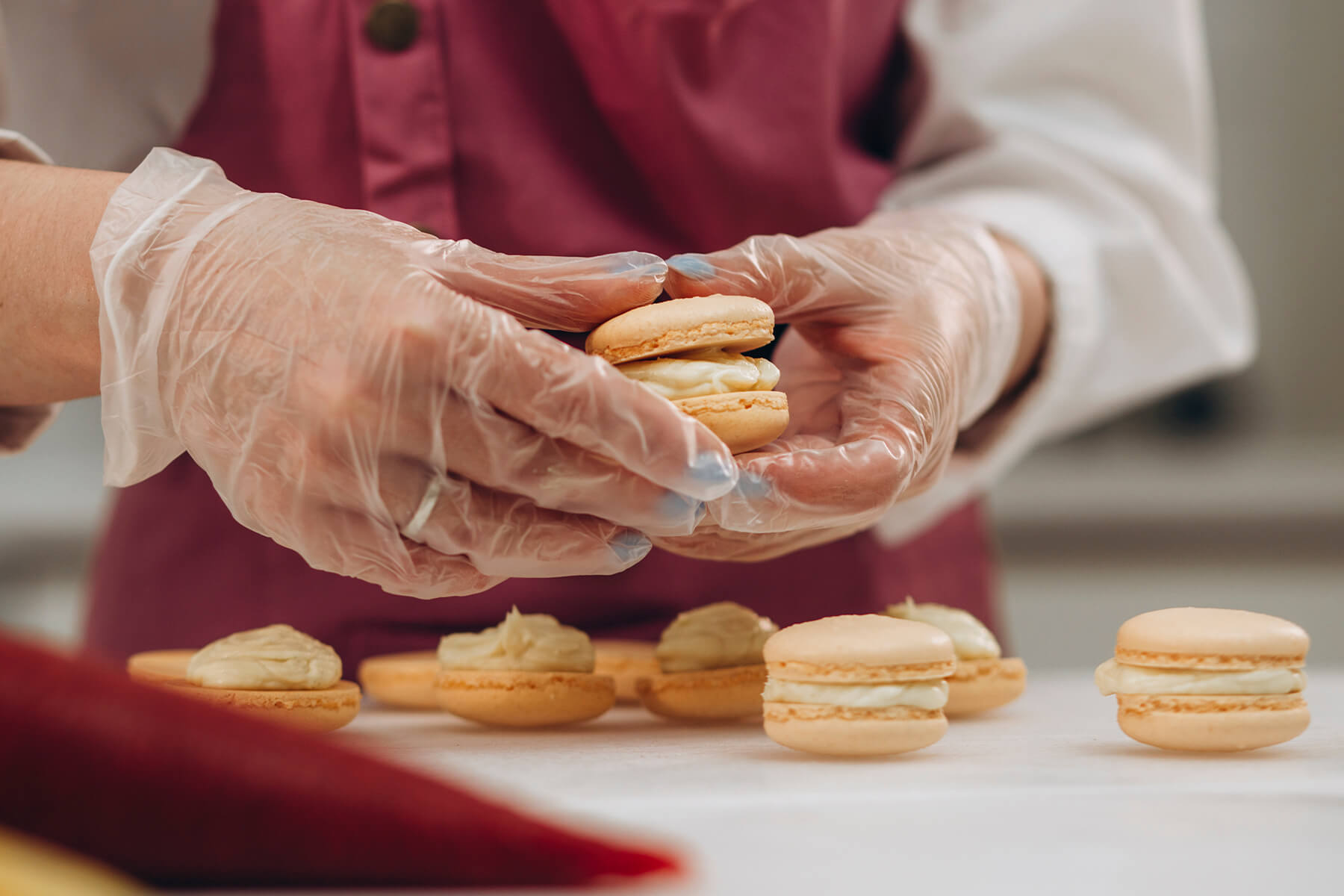 Behandschuhte Hände setzen zwei helle Macaronschalen zusammen, davor liegen weitere Schalen mit aufgespritzter Creme in einer Reihe